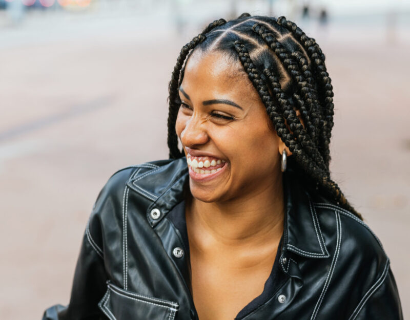Radiant woman with braided hair laughing in a casual outdoor setting. She looks happy enjoying the moment, wearing an stylish leather jacket.